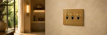 Solid brass three-gang toggle light switch with knurled brass levers placed on a beige wall in a contemporary home setting.