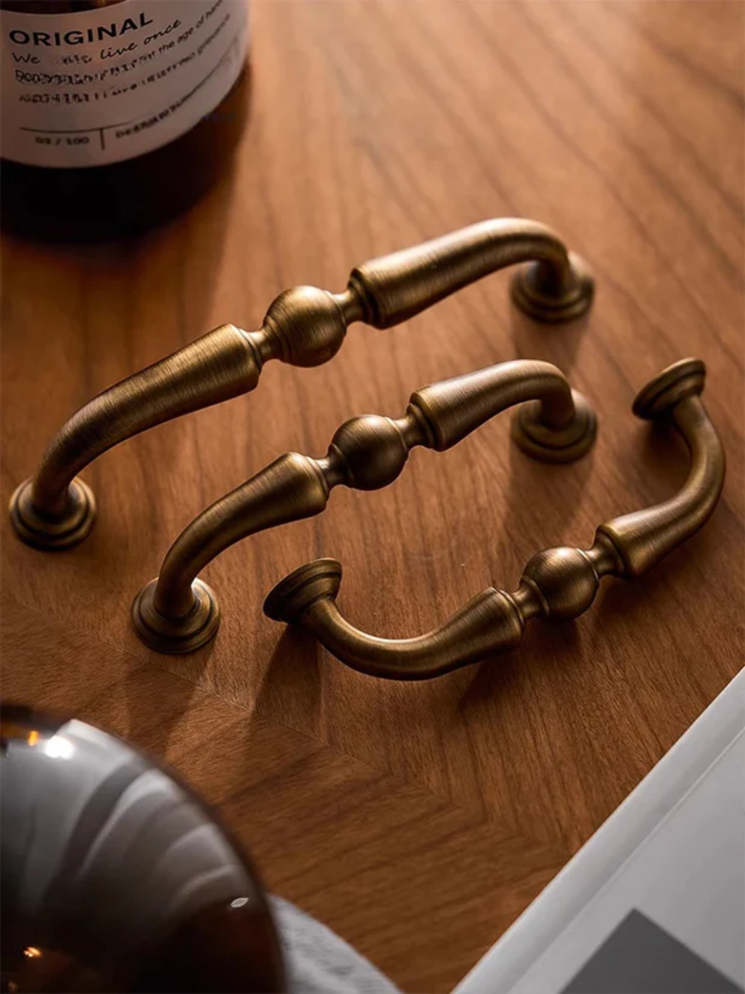 Two brass cabinet handles on a wooden surface with a bottle in the background.