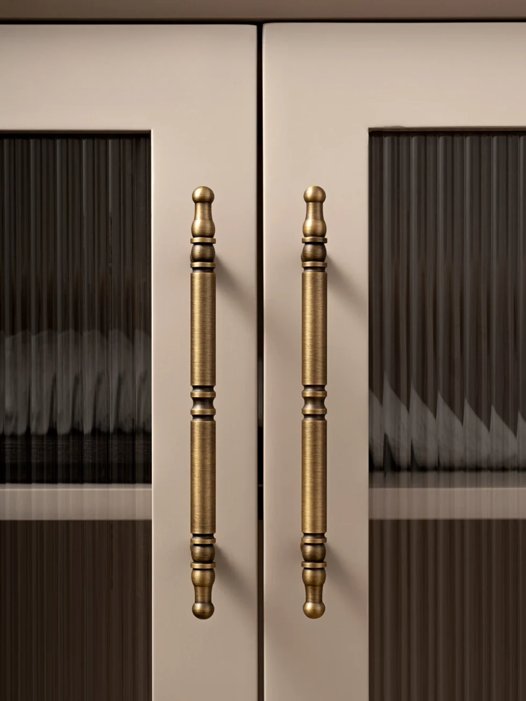 Close-up of a pair of bronze cabinet handles on a beige cabinet with dark glass panels.