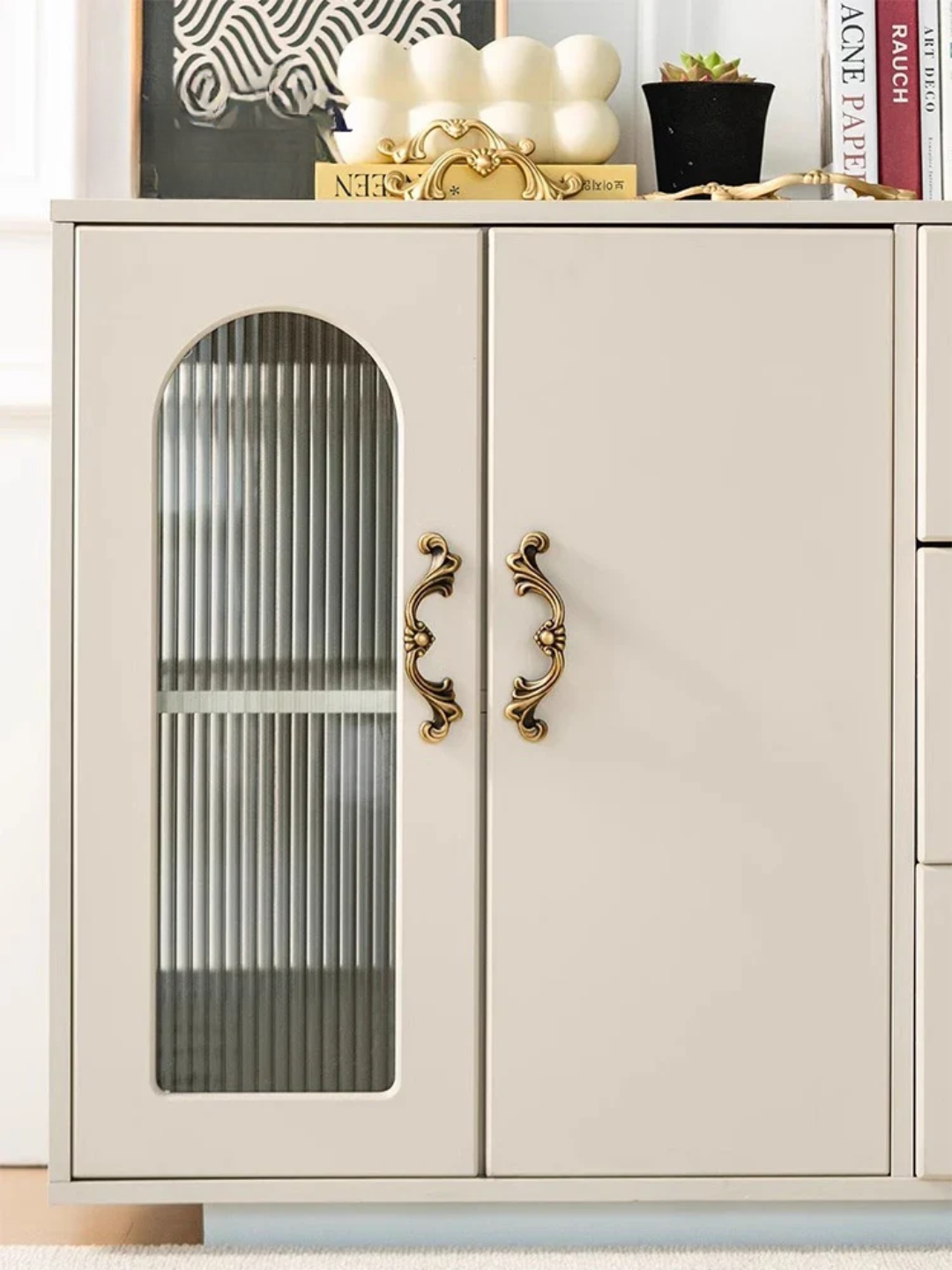 Beige cabinet with glass door and decorative handles, featuring a gold candle holder and books on top.