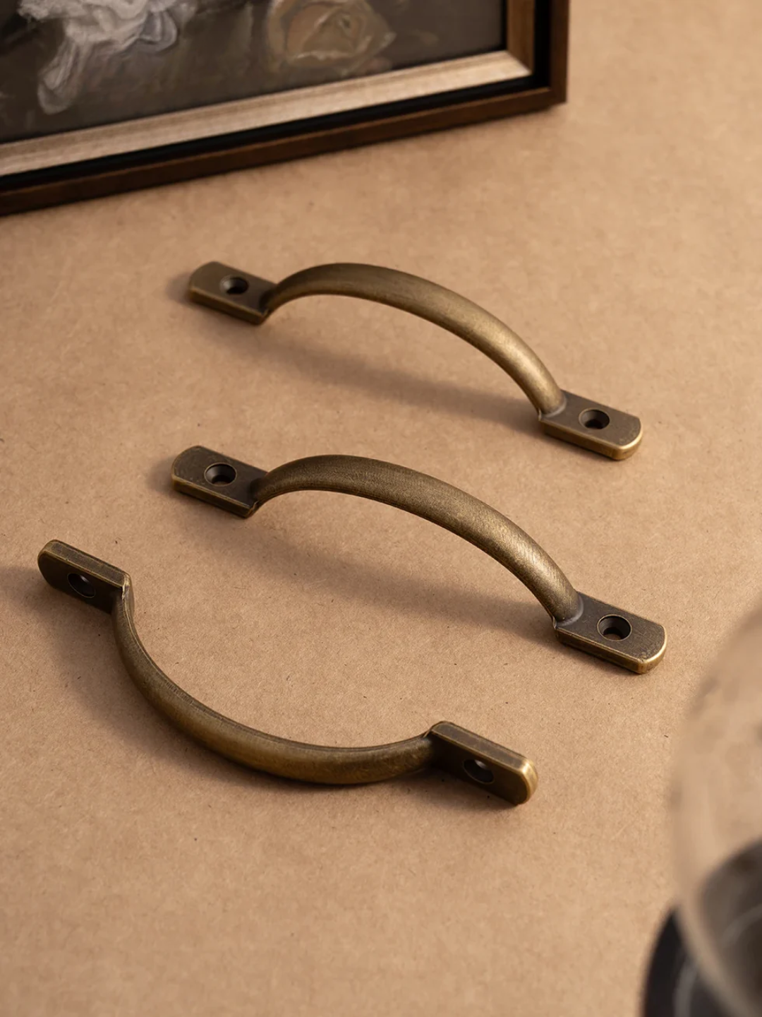 Two brass cabinet handles on a beige surface with a framed picture in the background.