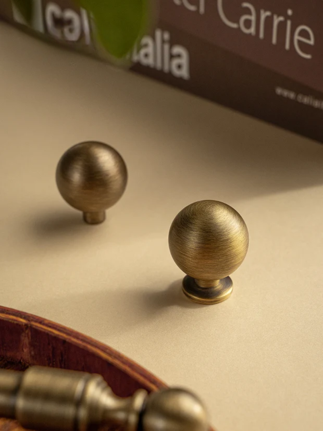 Two brass cabinet knobs on a beige surface with a blurred product box in the background.
