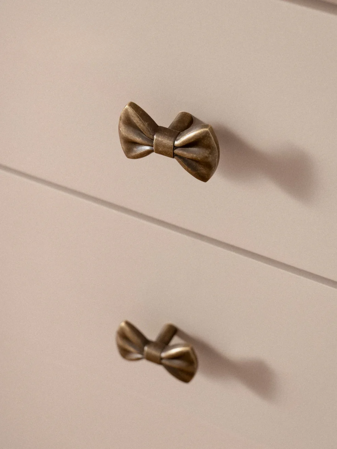Two bronze bow-shaped cabinet knobs on a beige drawer.