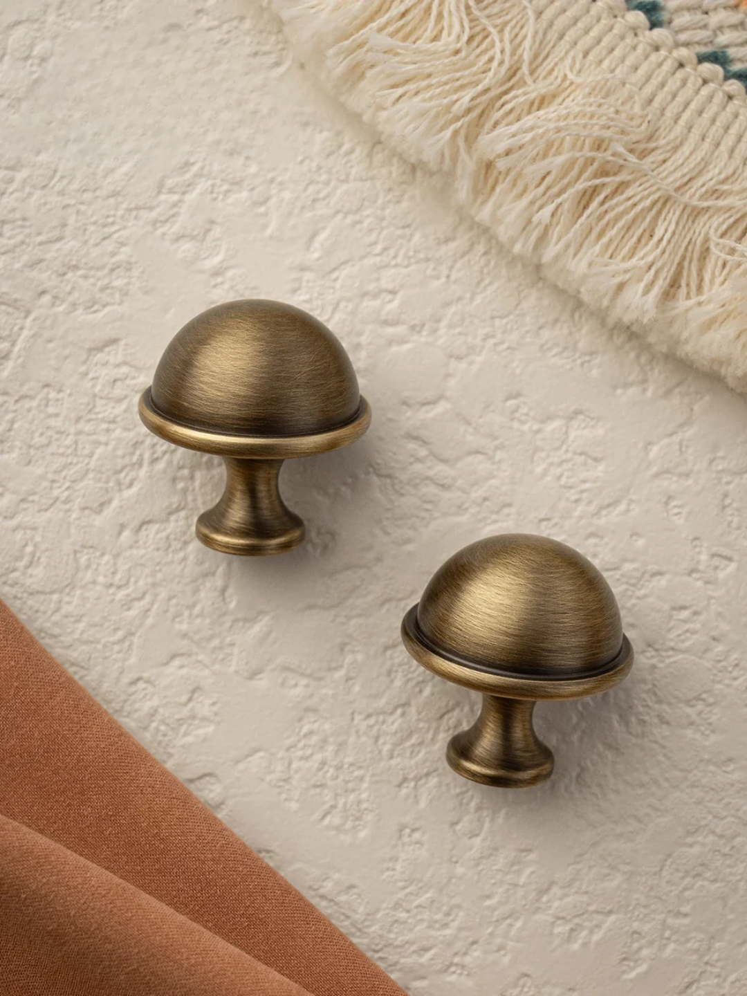 Two brass cabinet knobs on a textured beige surface with a fabric and rug in the background.