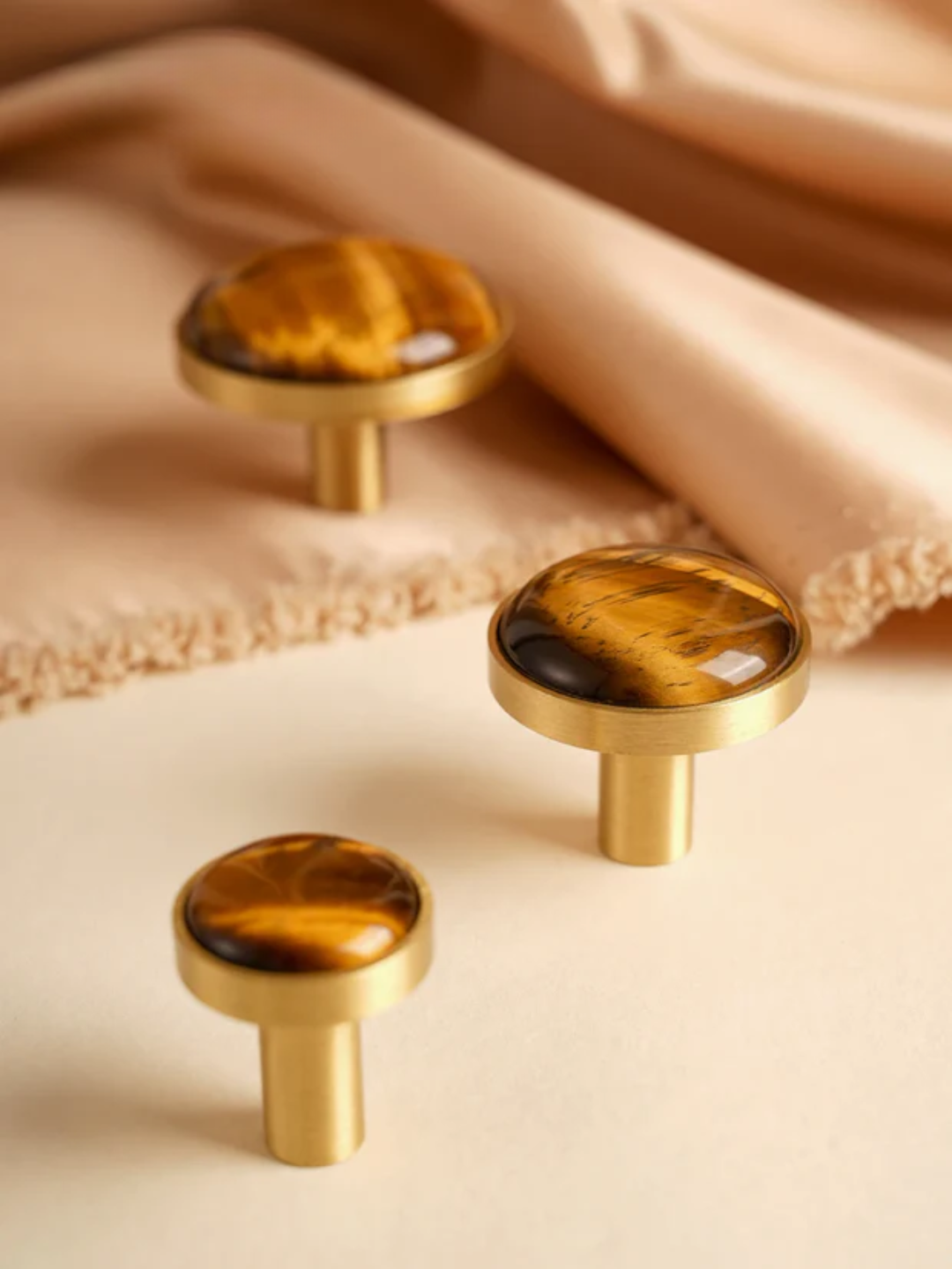 Three gold cabinet knobs with tiger eye stone tops on a beige fabric background
