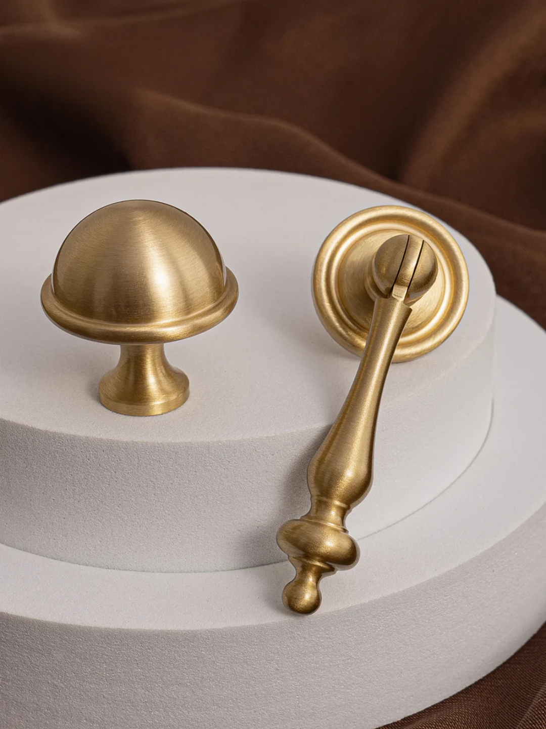 Two gold cabinet knobs on a white surface with a brown fabric background