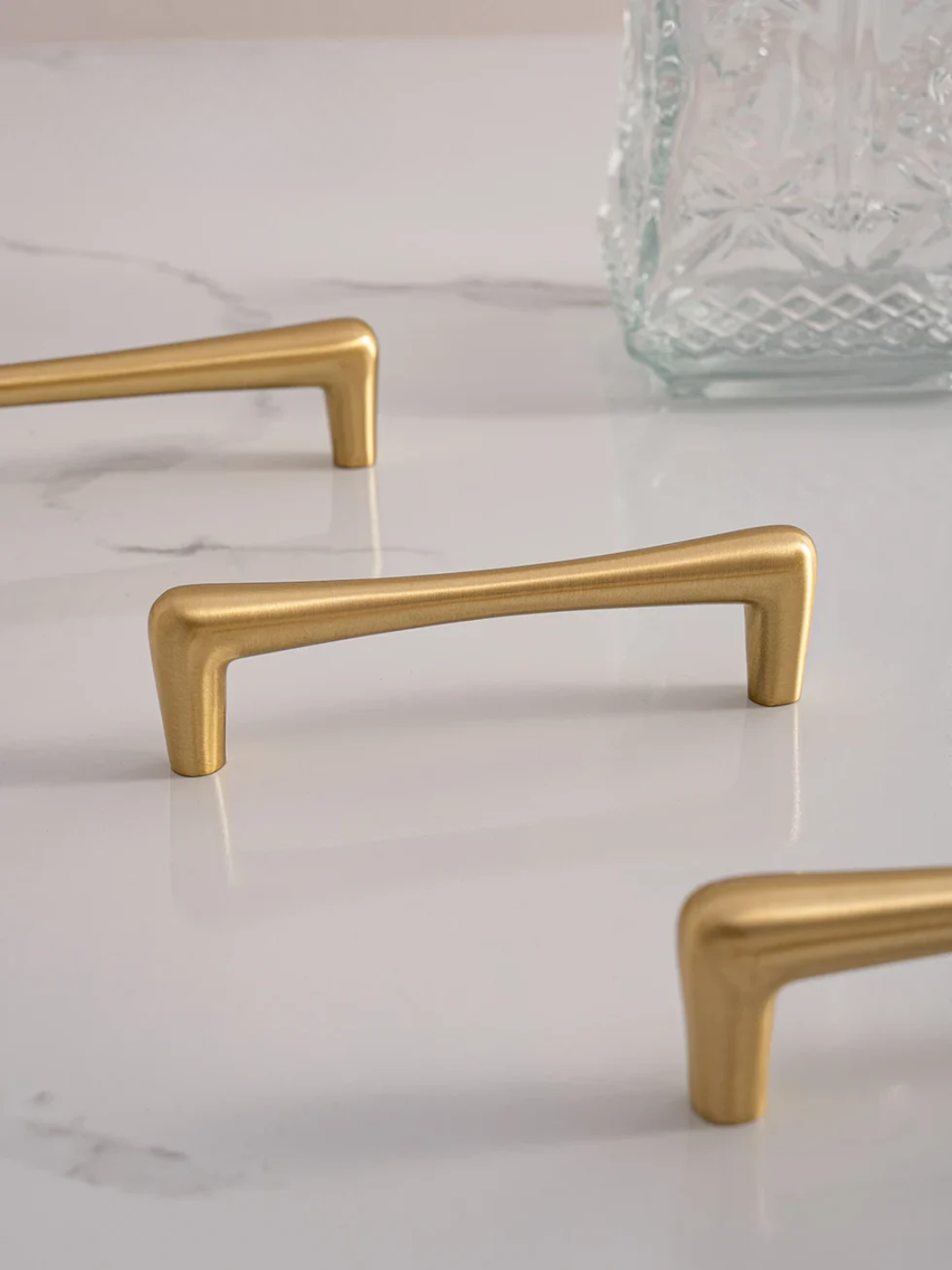Gold cabinet handles on a marble surface with a glass vase in the background.