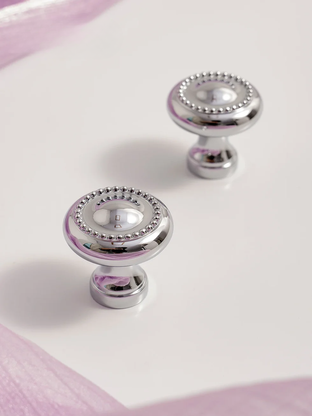 Two silver cabinet knobs with decorative beads on a white surface with pink edges.