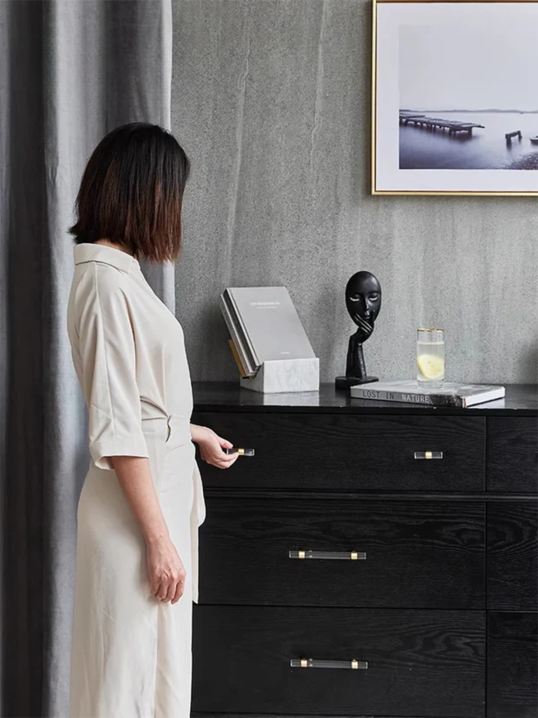 Woman standing in a room with a black dresser and decorative items.