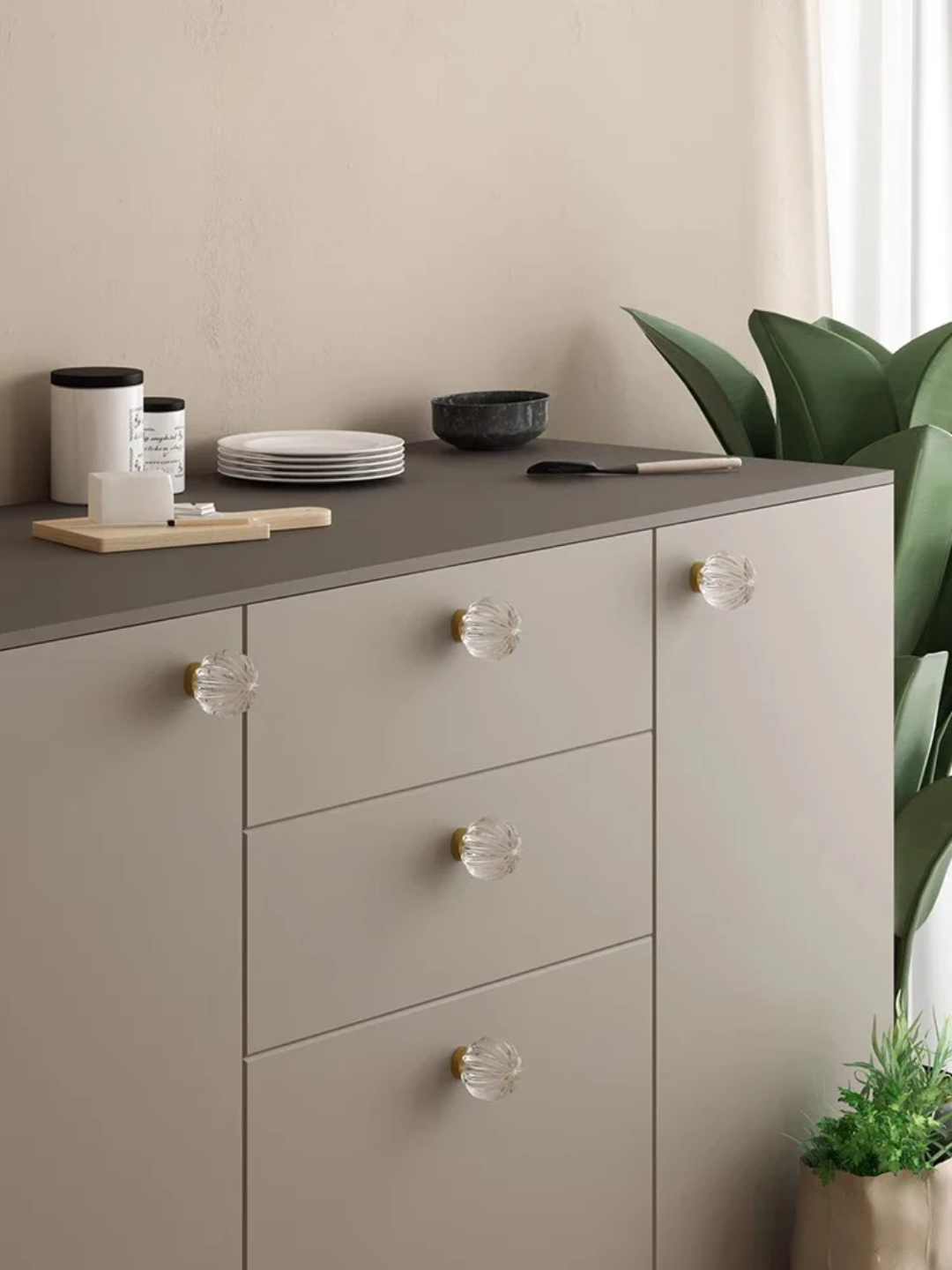 Gray dresser with clear glass knobs in a room with a plant and minimal decor.