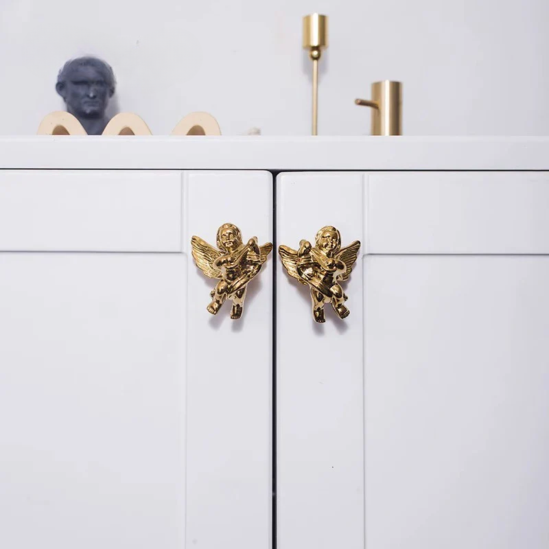 Gold angel-shaped cabinet handles on a white cabinet with decorative items above.