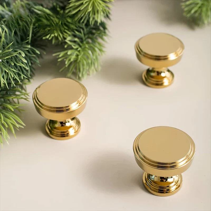 Three gold cabinet knobs on a light surface with green foliage in the background