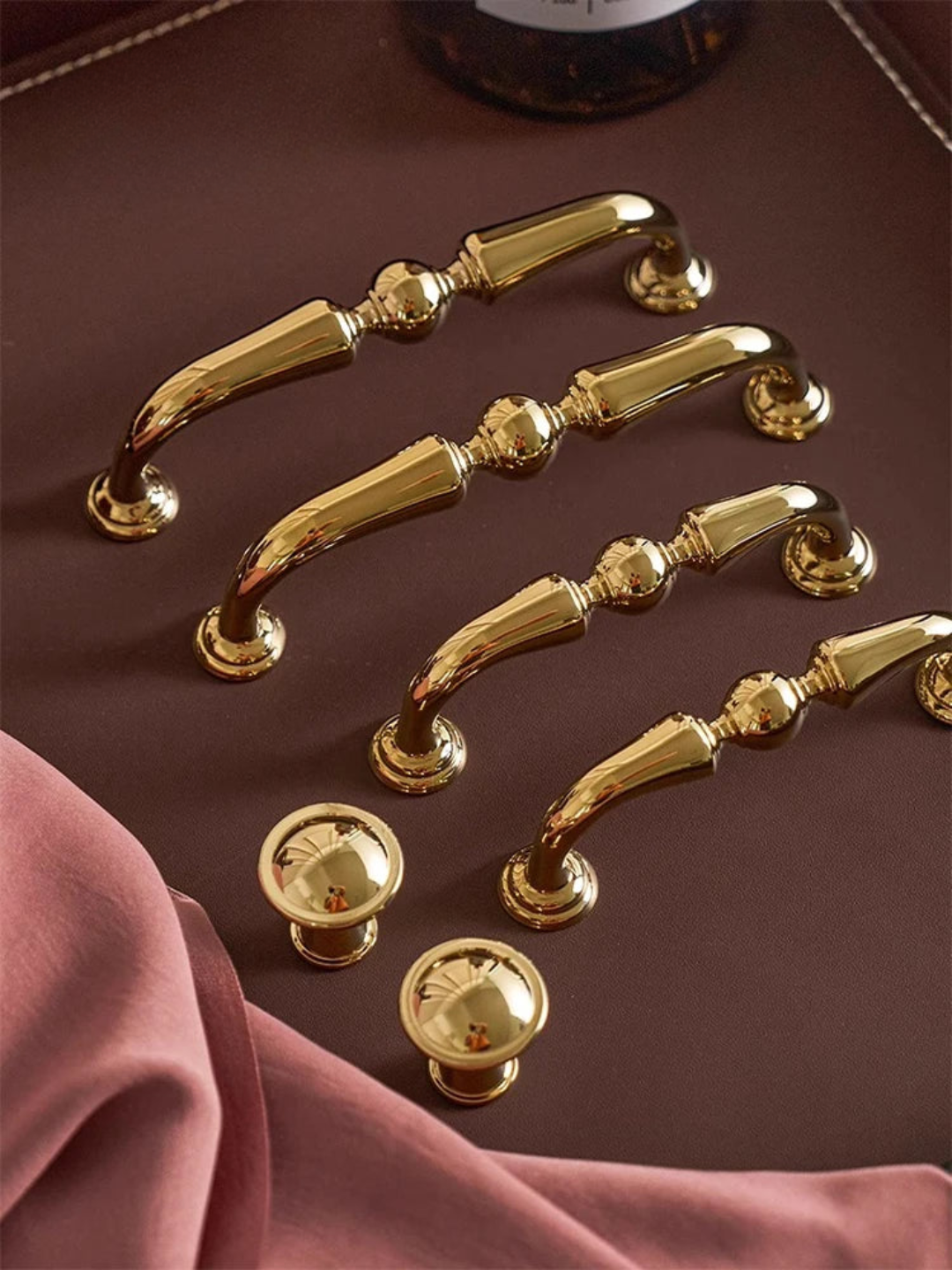 Set of gold cabinet handles and knobs on a brown surface with pink fabric.