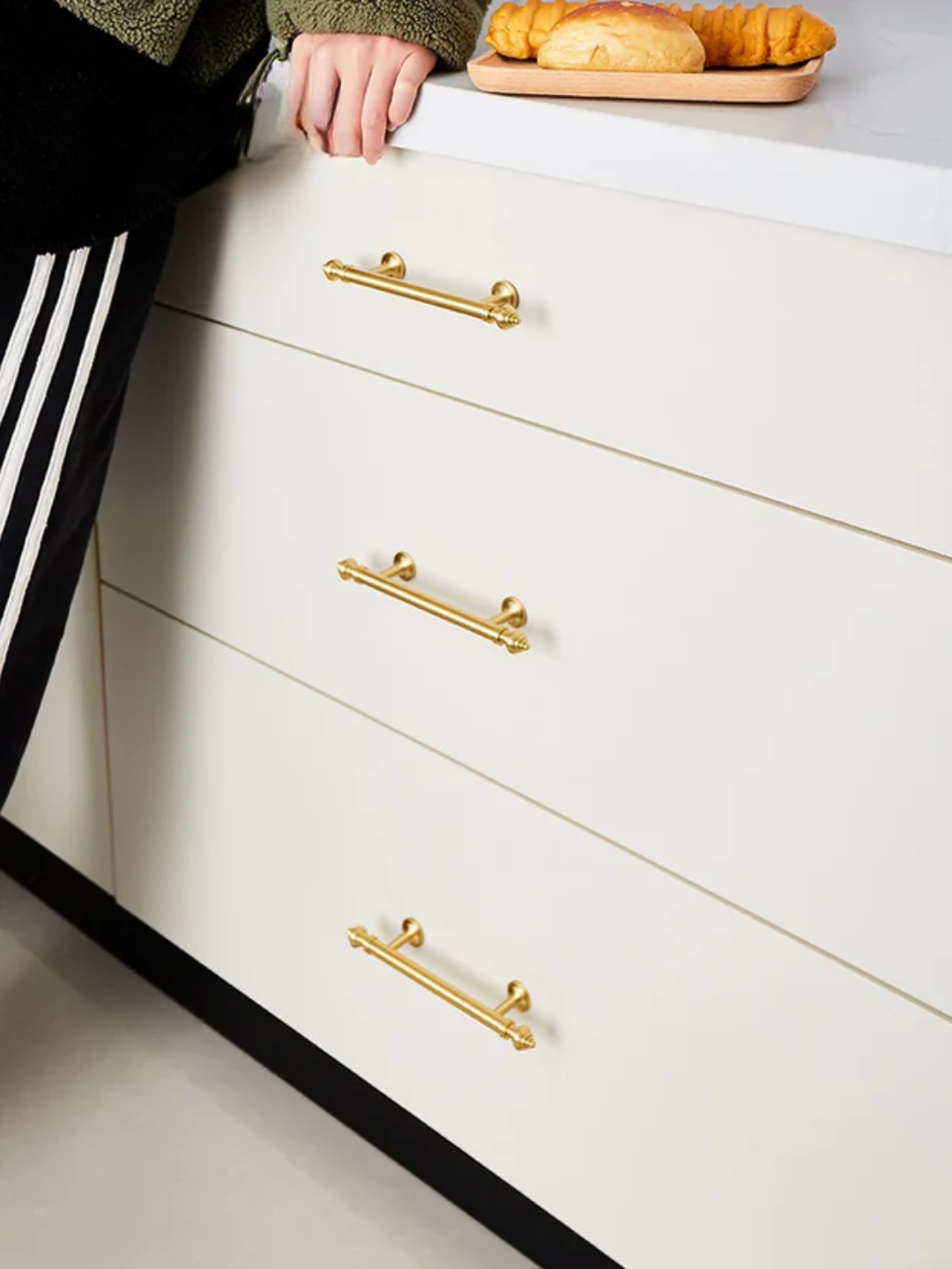 White kitchen cabinets with gold handles and a person wearing Adidas pants.