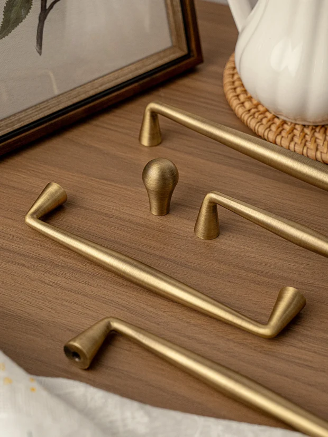Set of gold cabinet handles on a wooden surface with a teapot and framed picture in the background.