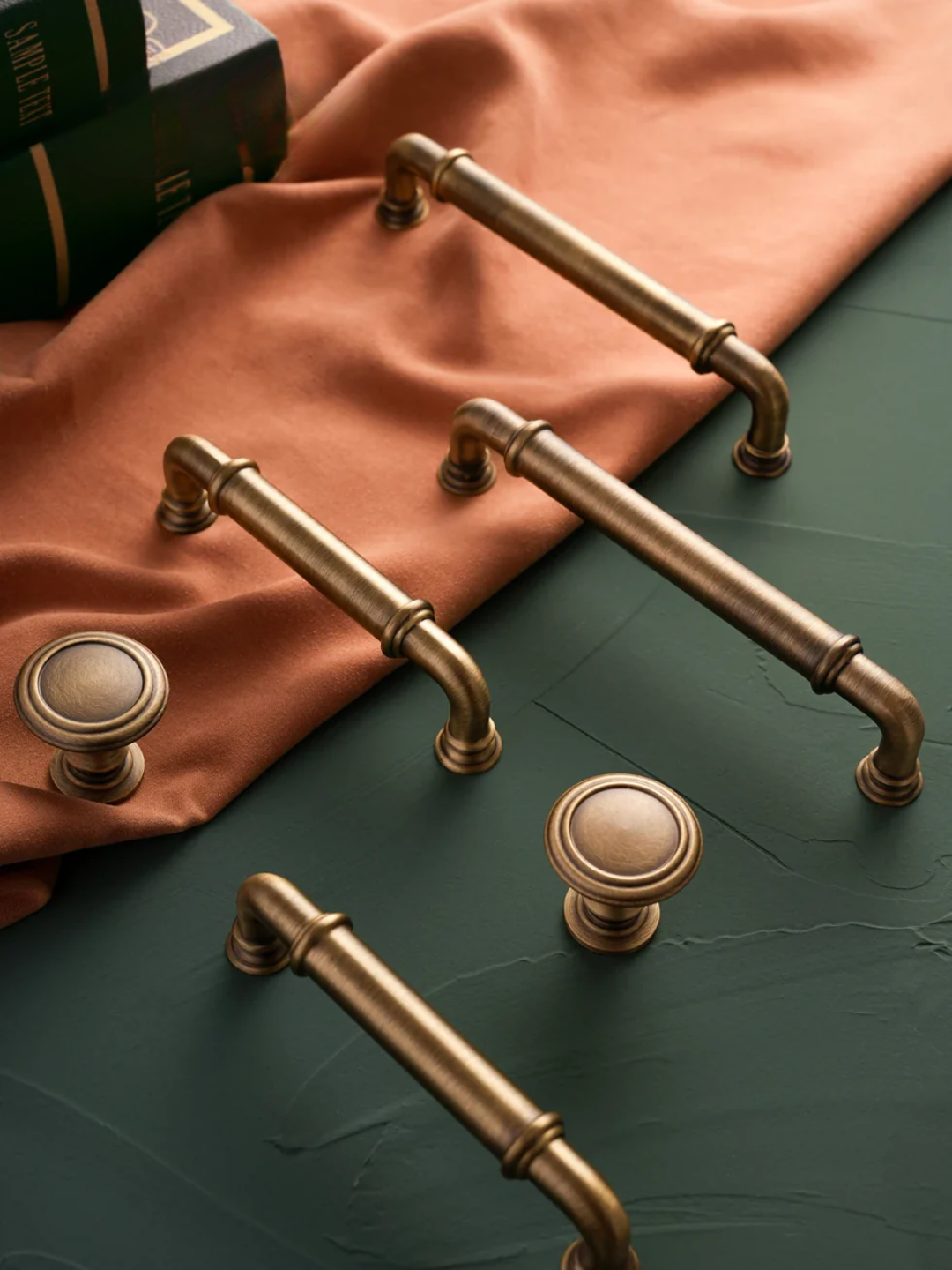 Bronze cabinet handles and knobs on a green surface with a rust-colored fabric background.