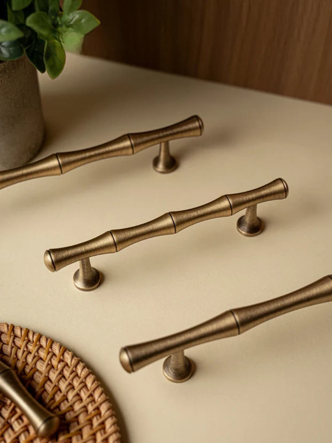 Set of bronze cabinet handles on a beige surface with a woven mat and plant in the background.