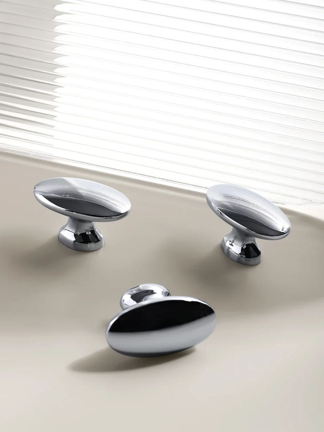 Three chrome cabinet knobs on a beige surface with white blinds in the background