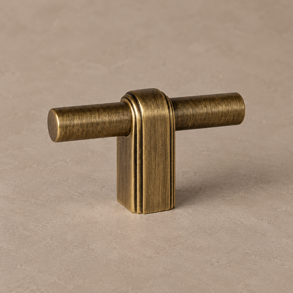 antique brass T knob, cylindrical horizontal bar through a rounded rectangular central post with parallel edge grooves on warm taupe surface