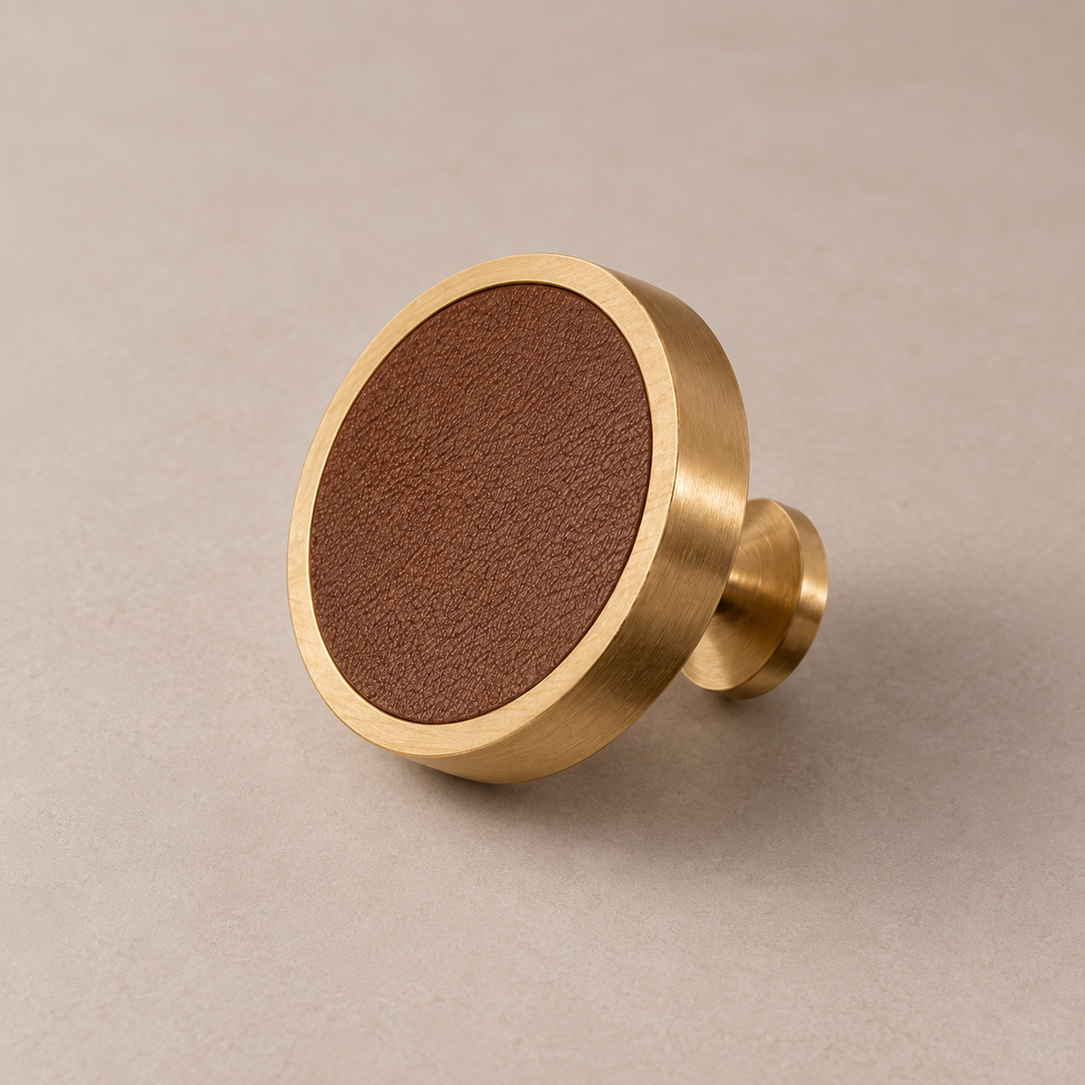 Brushed brass cabinet knob with brown cowhide leather inlay on taupe studio background