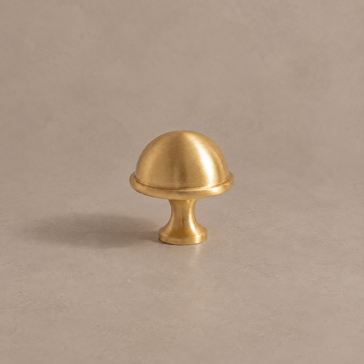 Brushed brass dome cabinet knob with circular rim and round base on a taupe studio background