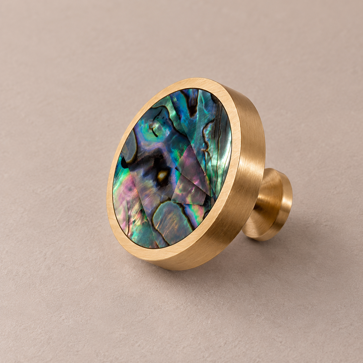 Brushed brass cabinet knob with green abalone shell inlay on taupe studio background