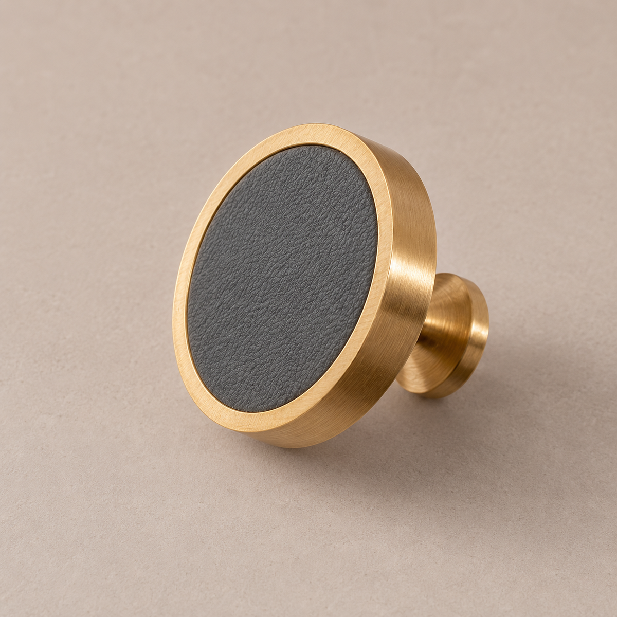 Brushed brass cabinet knob with grey cowhide leather inlay on taupe studio background