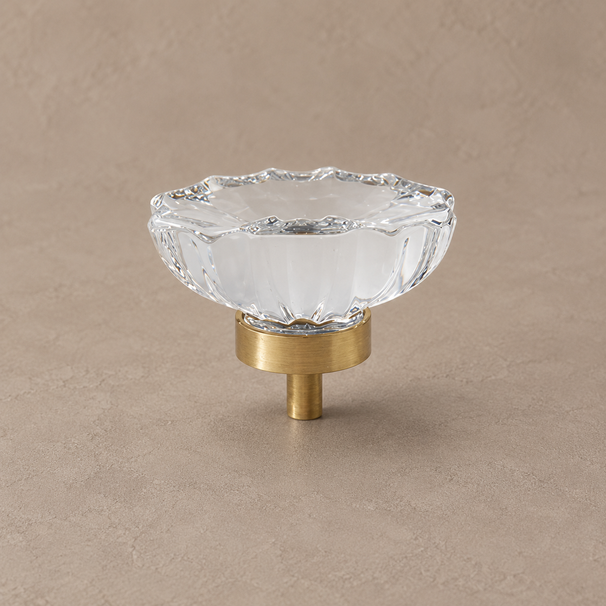 Clear scalloped crystal cabinet knob with brushed brass stem on warm taupe surface