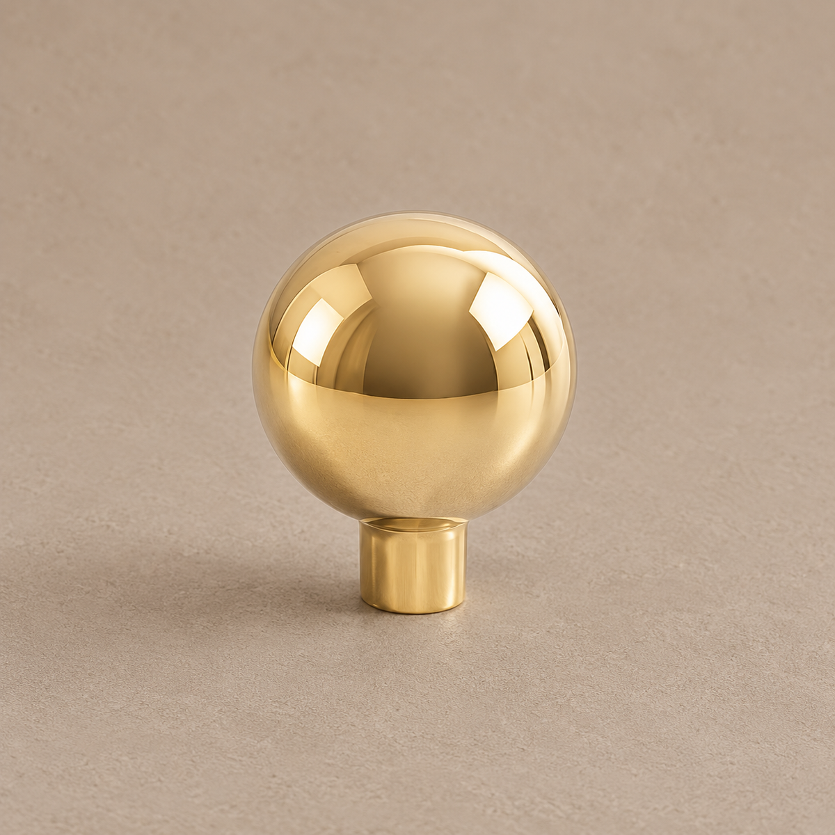 Small polished gold brass ball cabinet knob with a straight cylindrical stem on a taupe studio background
