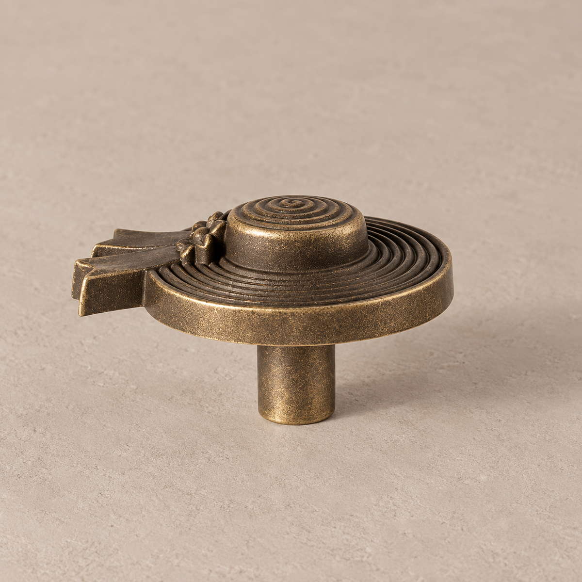 antique brass wide round hat/medal form cabinet knob with concentric grooves, small floral and ribbon ornament at one edge and smooth cylindrical mounting post on taupe studio surface