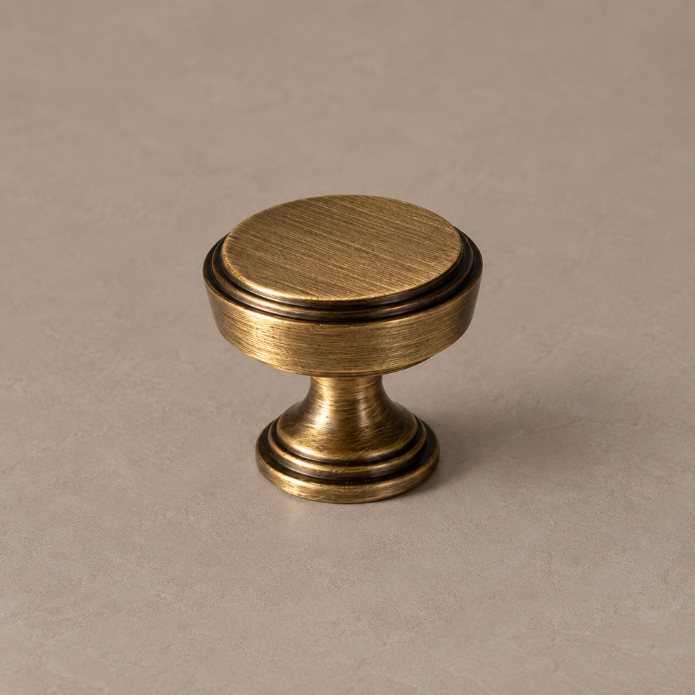 antique brass flat circular cabinet knob, stepped concentric rim, flared pedestal and circular stepped base on warm taupe surface